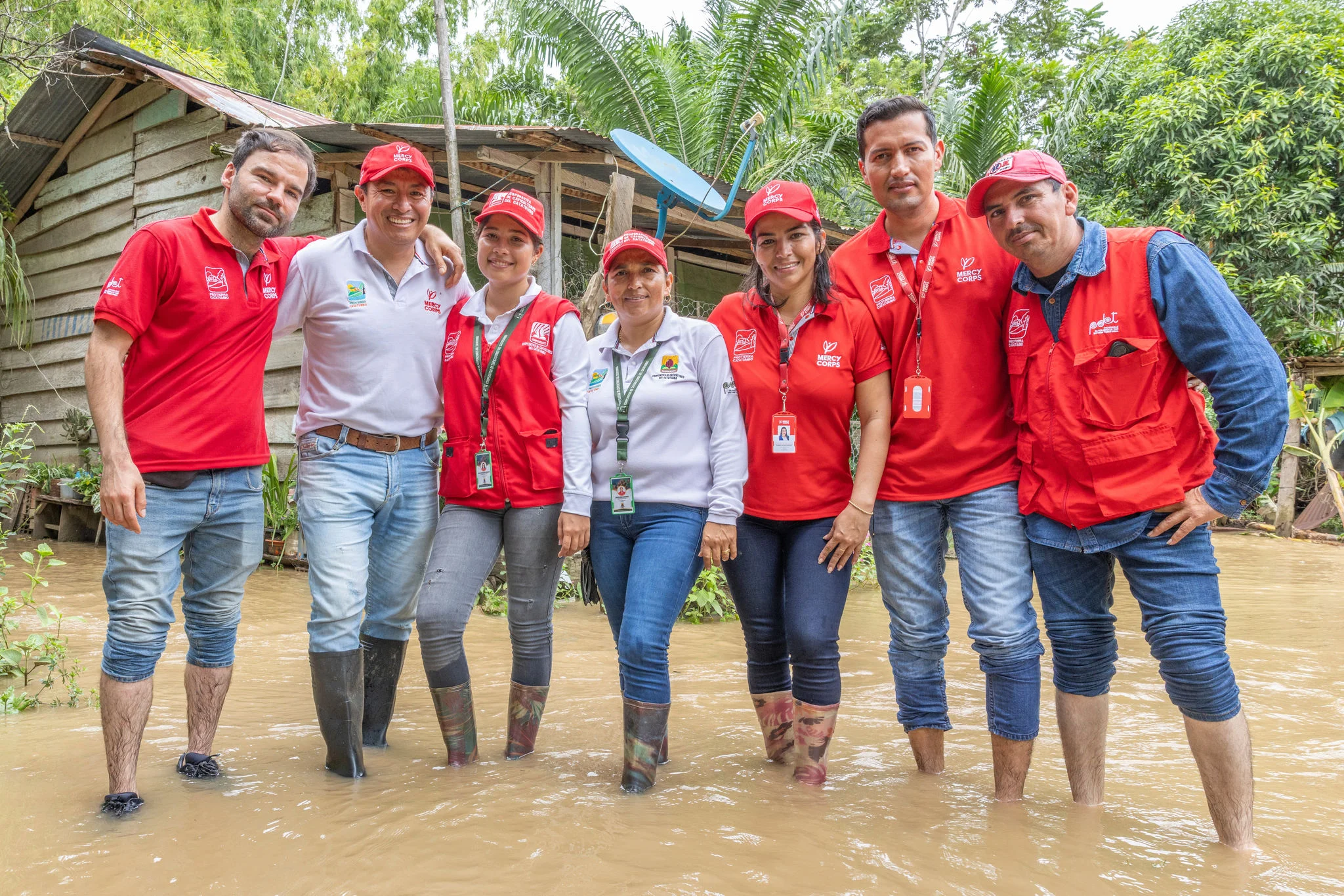 A group of seven relief workers wearing red and white Mercy Corps branded shirts and hats stand together in calf-deep floodwater in front of a modest wooden house.