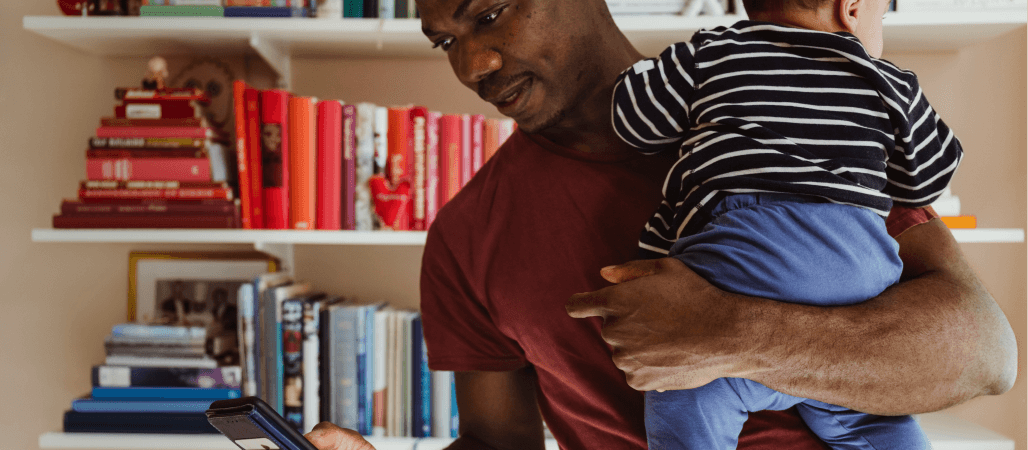 Image of man holding his child and looking at his phone while inside their home in front of a wall of books. 