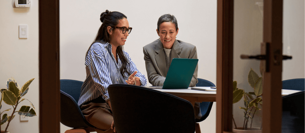 Image of two people sitting in an office space while looking at a laptop together.